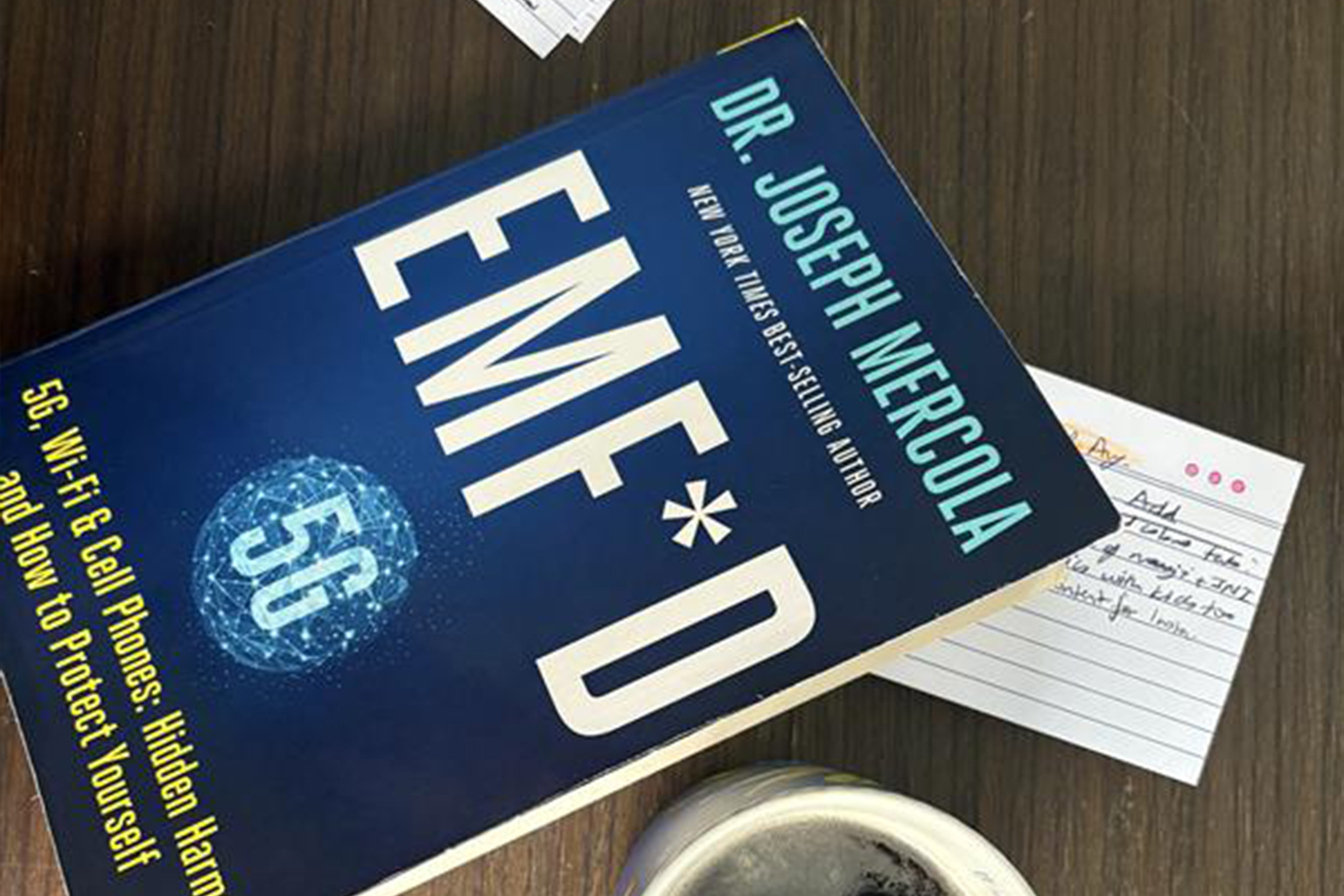 Book "EMF*D" by Dr. Joseph Mercola on a wooden desk with a cup of coffee and handwritten notes