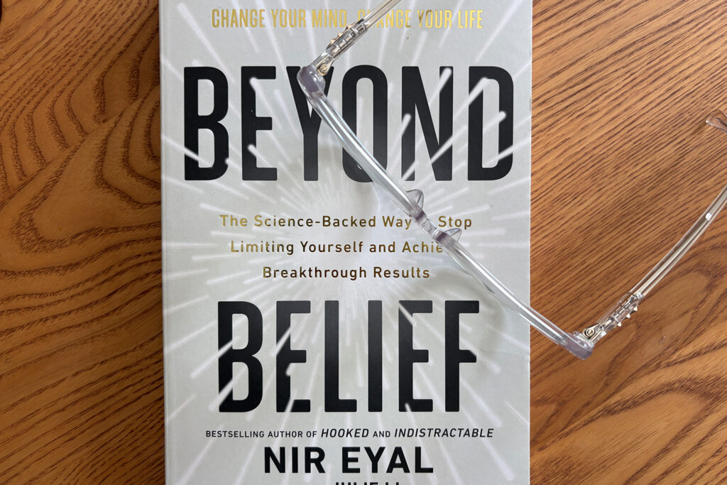 Copy of Beyond Belief by Nir Eyal on a wooden table with reading glasses, representing mindset shifts and behavior change